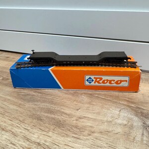 May include: A dark brown model railway flat wagon with black wheels and metal couplings, resting on a short section of track. The model is positioned on top of its original rectangular cardboard packaging, which features a blue and orange design with the Roco logo printed in white and blue.