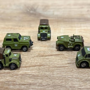 Pode incluir: Uma coleção de veículos militares em miniatura verde-oliva, incluindo caminhões, jipes e uma ambulância, dispostos sobre uma superfície de madeira. Cada veículo apresenta designs detalhados, incluindo estrelas e as letras "USMC".