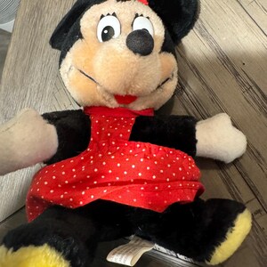 May include: A Minnie Mouse soft toy, featuring a red and white polka dot dress and bow. The doll has black ears, arms, and legs with yellow feet. The face is tan with black eyes and a red mouth. A tag is attached.