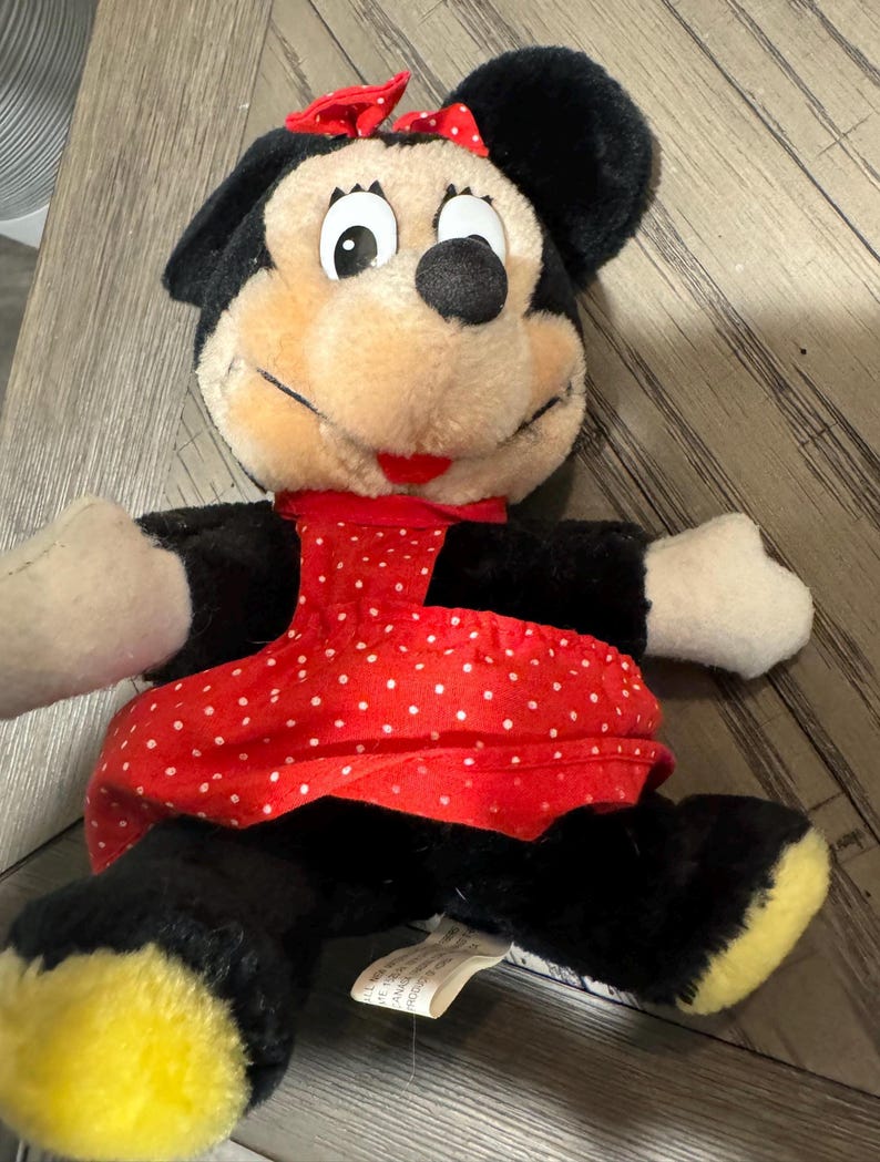 May include: A Minnie Mouse soft toy, featuring a red and white polka dot dress and bow. The doll has black ears, arms, and legs with yellow feet. The face is tan with black eyes and a red mouth. A tag is attached.