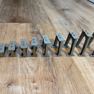 Puede incluir: Una colección de soportes arquitectónicos grises en miniatura, dispuestos en fila. Los soportes tienen una parte superior rectangular con un pequeño detalle en relieve y un diseño de pata arqueada. Los objetos están sobre un fondo de vetas de madera.