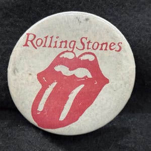 Op de afbeelding: Een ronde, crèmekleurige button met de rode tekst "Rolling Stones" en het iconische rode lippen- en tonglogo. Een klassiek muziekmemorabilia.