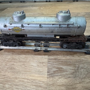 May include: Vintage silver-colored toy train tank car with two silver domes on top. The tank car has the Sunoco logo and text that reads "Built by Lionel". The train car is on a black metal chassis with black wheels and sits on a metal track.