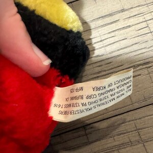 May include: A soft toy featuring red, black, and yellow colours. The toy is made from polyester fibres and has a tag stating "Product of Korea". The tag also includes the manufacturer's details.