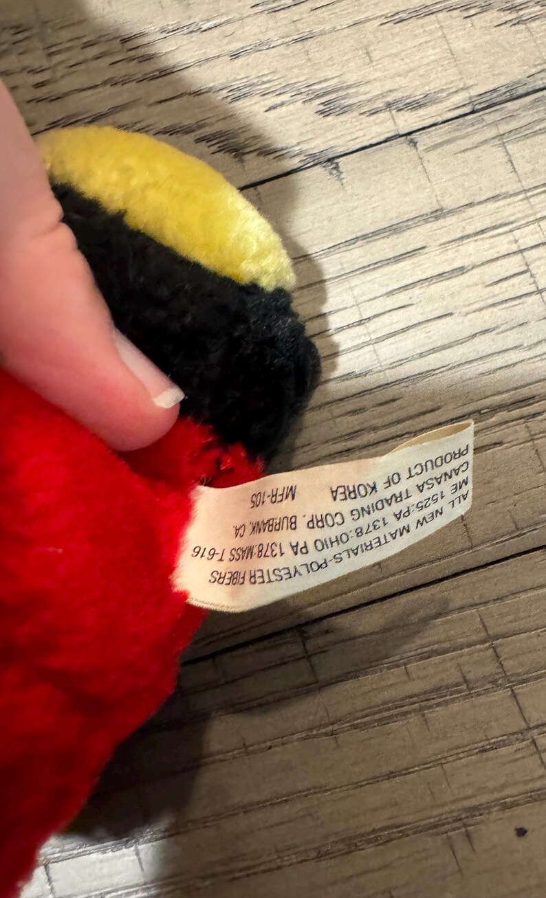 May include: A soft toy featuring red, black, and yellow colours. The toy is made from polyester fibres and has a tag stating "Product of Korea". The tag also includes the manufacturer's details.