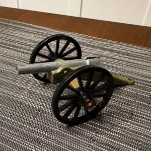Op de afbeelding: Een miniatuur kanonspeelgoed met zwarte spaakwielen en een zilveren loop. De behuizing van het kanon is olijfgroen en zilver. Het speelgoed staat tegen een gestructureerde grijze en bruine achtergrond.
