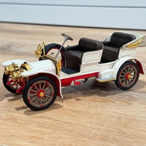 May include: A vintage-style white and red model car featuring dark brown tufted seats, gold-toned headlights, and a steering wheel. The vehicle has spoked wheels with red centres and a spare tyre mounted on the side, showcasing intricate detailing.