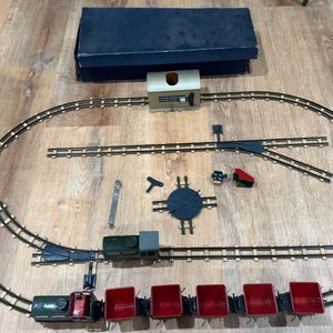 Könnte beinhalten: Ein Vintage-Modellbahn-Set mit ovalem Gleislayout. Das Set enthält eine dunkelgrüne Lokomotive, rote Güterwagen, einen Bahnhof und verschiedene Gleiskomponenten. Eine dunkelblaue Box ist oben.