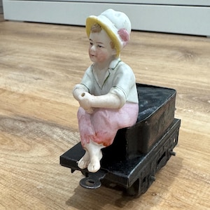 以下が含まれることがあります： 黄色い縁とピンクのアクセントが付いた白い帽子をかぶった子供のヴィンテージ磁器フィギュア。黒い金属製の玩具列車車に乗っています。子供は白いシャツとピンクのズボンを着ています。