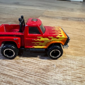 May include: A red toy pickup truck with black wheels and a black roll bar. The hood features a flame design in yellow and orange. The toy truck is a miniature replica of a classic vehicle.