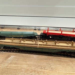 Puede incluir: Tres vagones de tren modelo sobre vías. Uno es verde con texto, otro rojo y el tercero marrón claro. Los vagones están sobre una superficie de madera. Son réplicas en miniatura de vagones de mercancías.