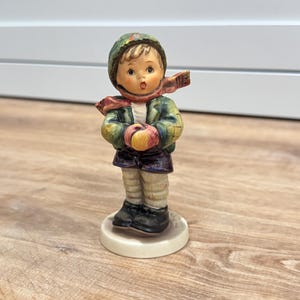 Op de afbeelding: Een keramieken beeldje van een kind met een groene muts, een groene jas, een rode sjaal, een donkerpaarse korte broek en een lichte legging. Het kind heeft lichtbruin haar, draagt zwarte schoenen en staat op een ronde witte voet.