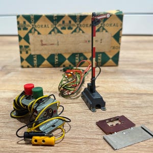 May include: A collection of model train accessories, including a red and white striped railroad crossing gate, control boxes with red and green knobs, and a box with the word "TROKAL" printed on it. The items are on a wooden surface.