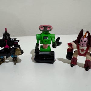 May include: Three colorful robot action figures. The first is black with gold and pink accents. The second is green with a pink face. The third is red, white, and maroon. They are on a white surface.
