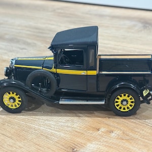 May include: Black and yellow vintage pickup truck model. The truck features a black body with yellow accents, including stripes and wheel details. The model has a spare tire and a wooden bed. The year 1929 is visible on the back.