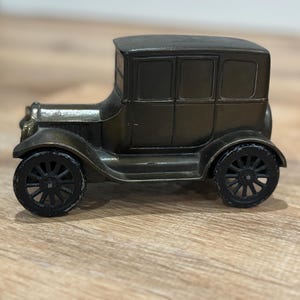May include: A bronze-colored antique car model with black wheels. The vintage vehicle has a classic design with a rectangular passenger compartment and a rounded roof. The wheels have a spoke design.