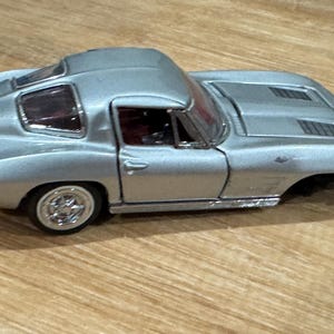 Op de afbeelding: Zilveren gegoten modelauto met een hardtop. De auto heeft een strak design met een gedetailleerd interieur zichtbaar door de ramen. De wielen hebben chromen velgen. Het is een klassieke sportwagen.