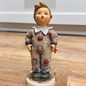 Puede incluir: Una figura de Hummel de un niño pequeño vestido de payaso, con una expresión de sorpresa. La figura tiene un atuendo de color púrpura claro y amarillo con detalles en rojo y amarillo. El niño está de pie sobre una base redonda de color blanquecino.