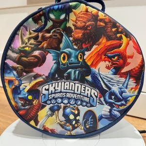 May include: Circular navy blue bag with a handle, showcasing characters from "Skylanders: Spyro's Adventure." The design is vibrant, featuring fantasy creatures and the game's logo. The bag is suitable for carrying items.