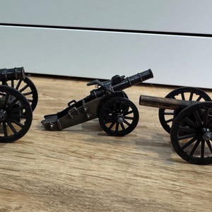 Op de afbeelding: Drie miniatuurkanonnen met zwarte wielen en donkere metalen lichamen worden getoond op een houten oppervlak. De kanonnen variëren in ontwerp, met één met een gebogen bovenkant en een andere met een langere loop. Het algemene thema is historisch.