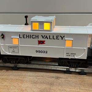 May include: A white Lehigh Valley caboose model with illuminated windows and a small roof structure. The model features the text "LEHIGH VALLEY" and the number "95032" on its side. The caboose is on a model train track.