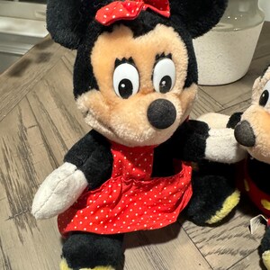 May include: A plush Minnie Mouse doll with black fur, a red polka-dot bow, and a red polka-dot dress. The doll has a beige face, large black eyes, and a black nose. The doll's feet are yellow.