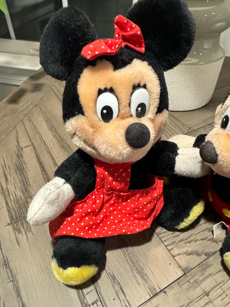 May include: A plush Minnie Mouse doll with black fur, a red polka-dot bow, and a red polka-dot dress. The doll has a beige face, large black eyes, and a black nose. The doll's feet are yellow.