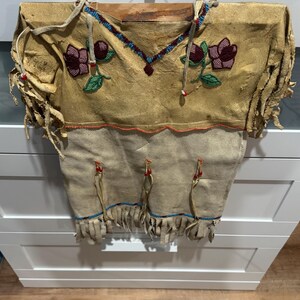 May include: A tan-colored, fringed garment with floral beadwork. The top portion features maroon and green beaded flowers, while the neckline and hem are adorned with blue and red beads. The garment is made of a soft, textured material.