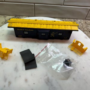 May include: A model train car from the Alaska Railroad, with a yellow roof and dark blue body. The text "ALASKA RAILROAD" and "6441" are visible. Includes yellow plastic parts, black pieces, and small parts in a bag.