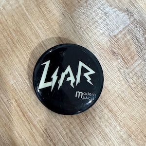 May include: A black button with the word "LIAR" in white, stylized, jagged lettering. Below the word "LIAR" is the text "modern music" in a smaller, white font. The button is circular and rests on a wooden surface.