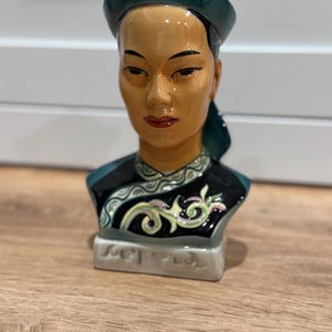May include: A ceramic bust depicting a woman with Asian features. She is wearing a teal hat and a black top with a decorative design. The bust has a tan complexion and red lips. The base is light grey.
