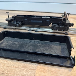 May include: Black toy train car with a flatbed design, sitting on a metal track. A separate black tray is in the foreground. The train car has small wheels and detailed features. The scene is set on a wooden surface.