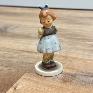 Könnte beinhalten: Eine Keramikfigur einer jungen Person mit braunem Haar, das mit einer blauen Schleife gebunden ist. Sie trägt ein dunkelbraunes Oberteil und ein hellblaues Kleid mit kleinen roten kreisförmigen Mustern, beigen Socken und dunkelbraunen Schuhen.