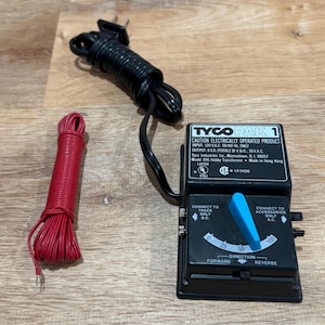 May include: A black Tyco Pak 1 model train transformer with a blue control lever, a black power cord, and a red wire bundle. The transformer has text that reads "CAUTION ELECTRICALLY OPERATED PRODUCT".