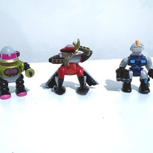 May include: Three toy robots in a row. The first is gold and pink, the second is red and gold, and the third is gray and blue. Each robot has a unique design and pose, suggesting a theme of action figures or collectibles.
