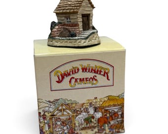 Può includere: Una statuetta in miniatura di un cottage in resina con tetto a scandole marroni e base in pietra, appoggiata su una scatola di cartone quadrata con l'etichetta David Winter Cameos, raffigurante una scena di mercato di villaggio con persone e cavalli.