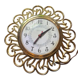 Pode incluir: Um relógio de parede dourado com uma moldura decorativa e ornamentada. O mostrador do relógio é branco com marcadores de hora em algarismos romanos pretos e ponteiros pretos. O mostrador do relógio também tem a palavra "UNITED" impressa.