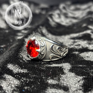 May include: A silver ring with a large, oval, red gemstone. The ring has an ornate, engraved design on the band and around the gemstone. The image also includes the text "NK RINGS" in a stylized font.
