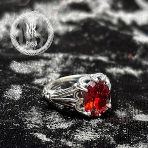 Może przedstawiać: Srebrny pierścionek z owalnym, ciemnoczerwonym kamieniem szlachetnym. Pierścionek ma bogaty, gotycki design ze skomplikowanymi detalami wokół kamienia. Logo "NK RINGS" znajduje się w okręgu w lewym górnym rogu.
