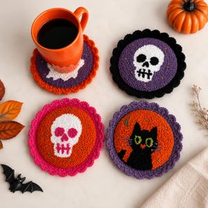 Pode incluir: Quatro porta-copos redondos de punch needle com bordas recortadas. Um apresenta uma caveira branca sobre roxo, um uma caveira branca sobre laranja, um gato preto sobre laranja e uma forma de fantasma branca sobre roxo sob uma caneca.