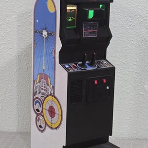 Puede incluir: Una máquina recreativa negra en miniatura con un panel lateral colorido con un gráfico retro con temática espacial. El juego tiene una pantalla, joysticks y el texto "BATTLEZONE" en la parte superior. El mueble mide aproximadamente 25 cm de altura.