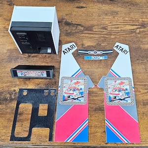 Puede incluir: Un conjunto de piezas de juego de arcade Atari, que incluye un gabinete, un panel de control y arte lateral. El arte lateral presenta un diseño de rayas de carreras rojo, blanco y azul con la palabra "ATARI" impresa en negro. El panel de control es negro con texto blanco e incluye botones para "CREDIT" y "START".