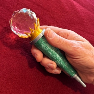 May include: A decorative wand with a faceted crystal orb, gold crown detail, and a green textured handle. The wand has a silver tip and is held in a hand against a red background. The wand is approximately 10 inches long.