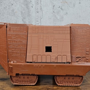 Könnte beinhalten: Ein braunes Plastikspielzeugmodell eines Star Wars Imperialen Truppentransporters, auch bekannt als TIE-Bomber. Das Modell hat ein detailliertes Äußeres mit einer strukturierten Oberfläche und einer kleinen Öffnung an der Seite. Das Modell hat einen Satz Räder an der Unterseite.