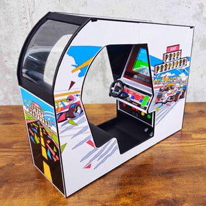 Puede incluir: Un gabinete de arcade blanco y negro con una cubierta de plástico transparente. El gabinete tiene un tema de juego de carreras con un diseño colorido que presenta un coche de carreras, una bandera a cuadros y el texto "Pole Position".