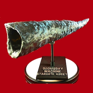 May include: A silver and blue metallic sculpture of a futuristic weapon, resembling a large, curved tube. The sculpture is mounted on a black stand with the text "DOOMSDAY MACHINE STARDATE 4202.1" on the base.