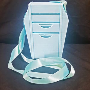 May include: A light blue, three-drawer box with a matching ribbon. The box has a rectangular shape with a slightly tapered design. The ribbon is looped and lies in front of the box, creating a decorative element.