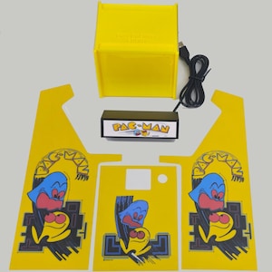 May include: A yellow Pac-Man arcade cabinet kit with a black control panel and a yellow Pac-Man character on the side panels. The kit includes a USB cable and a black box with the text "PAC-MAN" on it.
