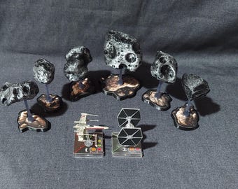 May include: A set of six miniature grey and black asteroid bases for a tabletop game. The bases are made of plastic and feature a textured surface. Each base has a small, clear plastic stand that allows the asteroid to be placed on a table or other surface. The bases are designed to be used with miniature spaceships or other game pieces.