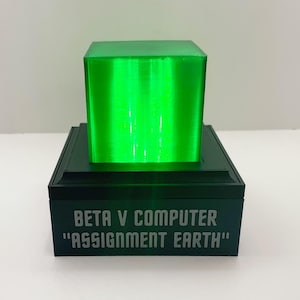 May include: A black box with the text "BETA V COMPUTER "ASSIGNMENT EARTH"" on the front. The box has a green, translucent cube on top.